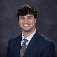 Studio headshot of a smiling person wearing a navy suit and patterned tie against a dark gray background.
