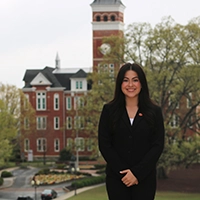 Professional portrait of a woman in business attire, standing outdoors with a historic university building in the background.