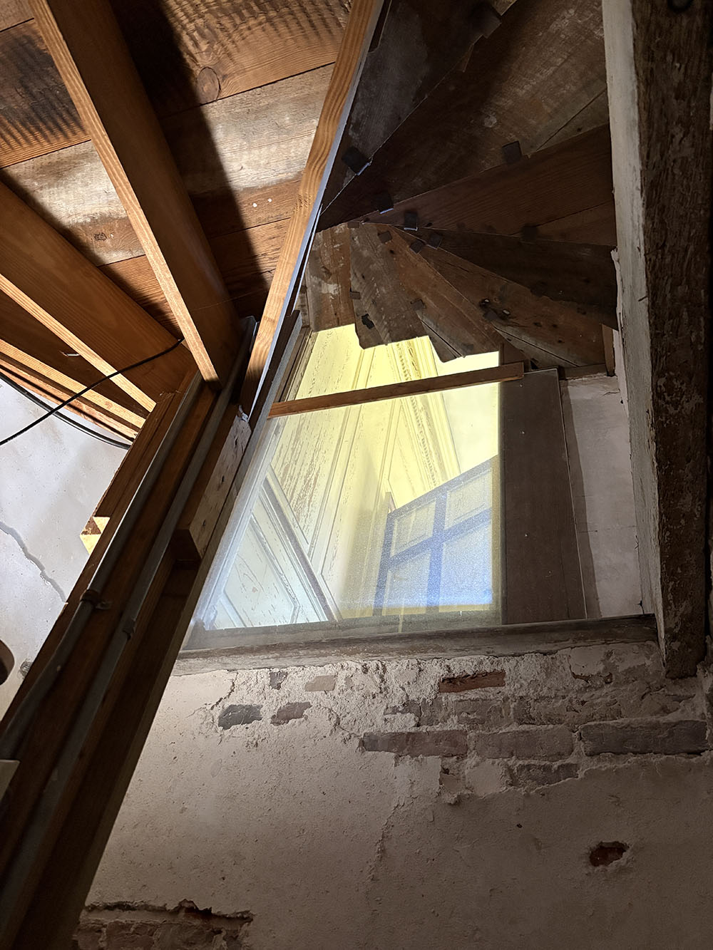 Upward view of exposed wooden beams and a sloped roof, with a small window letting in light above a rough plaster-and-brick wall.