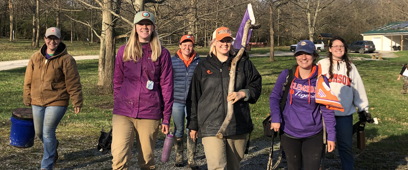 group of smiling students walking across a grassy field carrying buckets and digging equipment