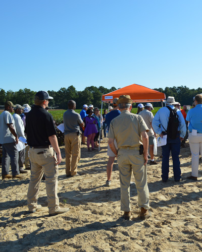 a crowd of people standing in a dirt roadway beside row crops