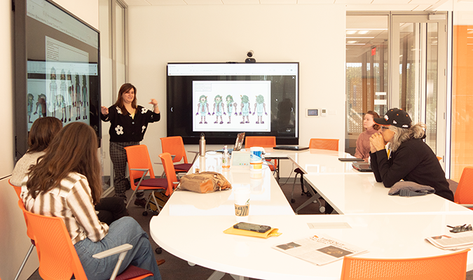 A young woman presents animated images on a screen in a classroom. The classroom is well-lit with large windows, a white table, and orange chairs.