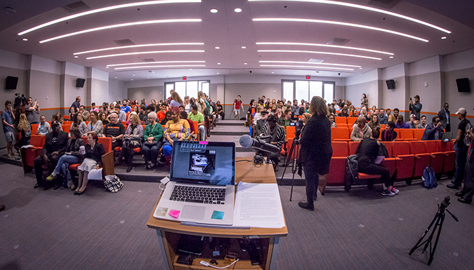 An image of a podium and a microphone from the speaker's perspective in front of a crowded room