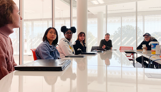 Students and faculty speak at a table in a classroom with large windows