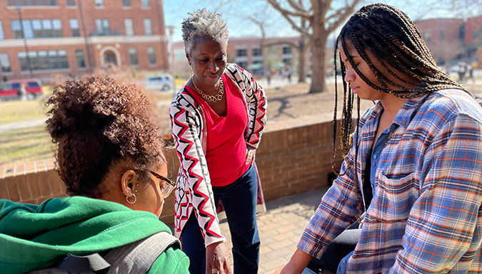 Three women gather outdoors and discuss a book