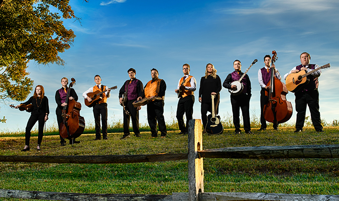 Bluegrass ensemble stands by fence on hill holding instruments
