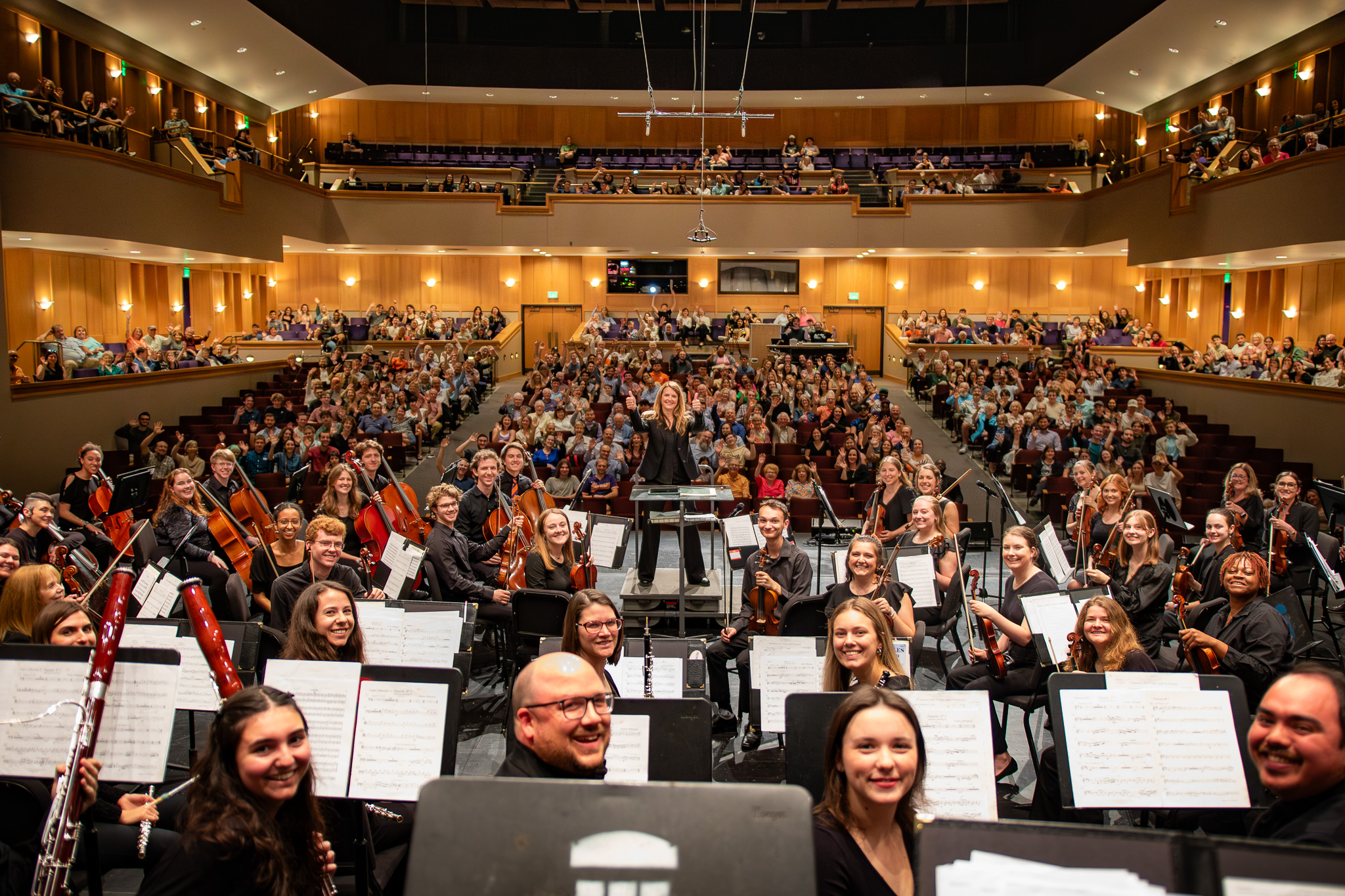The CU Symphony Orchestra smiles for a selfie on stage with the Brooks Center audience waving in the background