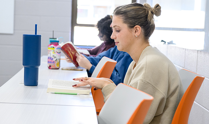 A student sits at a desk in class smiling while reading.