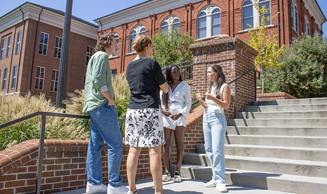A group of students talk with a professor outside Hardin Hall, a large brick building