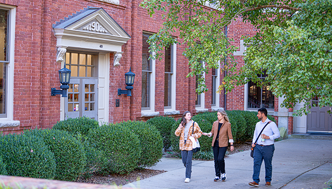students walk and talk with a faculty member on campus in front of a brick building