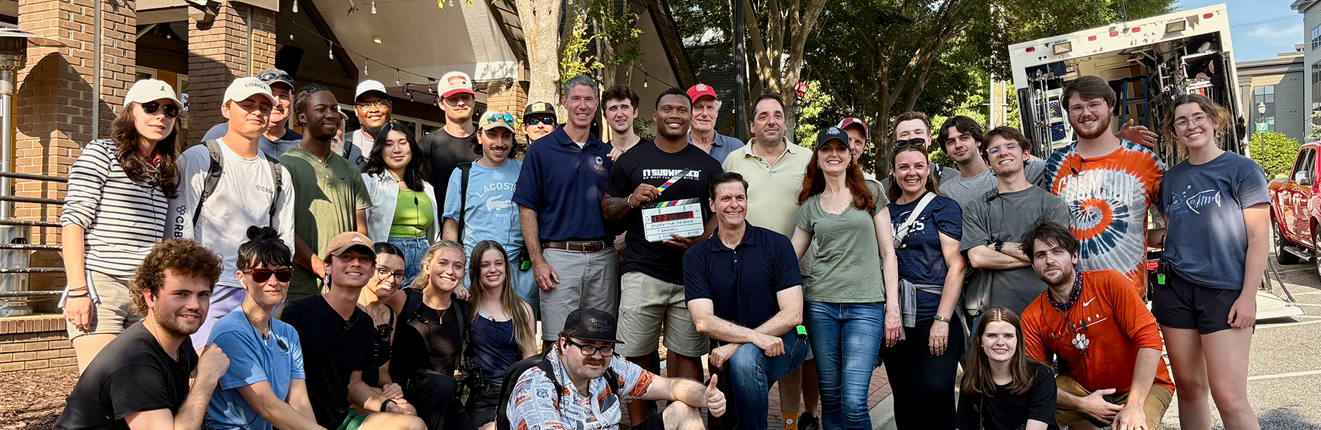 A group of people smiling on a sunlit street. One holds a clapperboard.