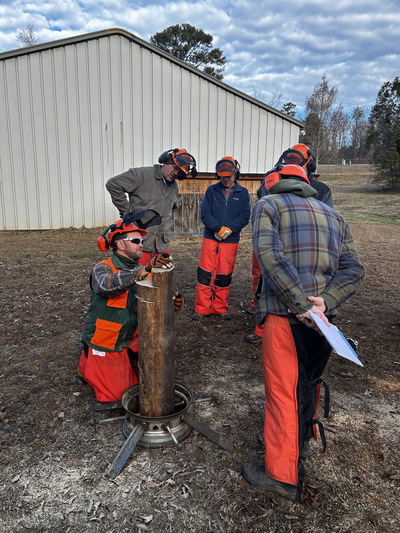 Group of workers wearing chainsaw safety helmets and chaps gather outdoors as one kneels to demonstrate on a vertical log set in a metal stand beside a shed.