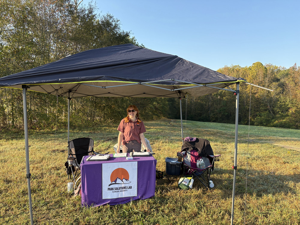 Outdoor booth under a pop-up canopy in a grassy field, Lydia Heisel standing behind a table covered by a Park Solutions Lab Clemson University banner.