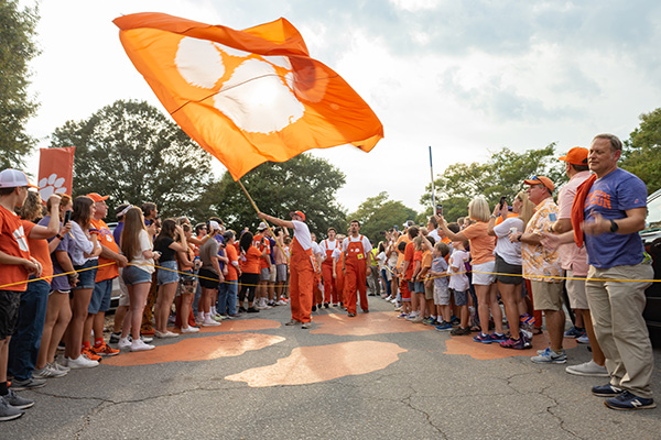 Man in orange overalls waving a large orange flag with a white Clemson tiger paw between fans on each side of a road.