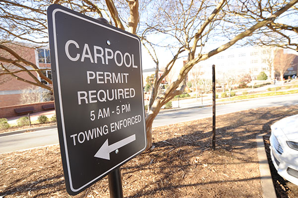 Brown sign with Carpool permit required 5AM to 5PM Towing enforced and an arrow pointing to the left in front of a leafless tree. 