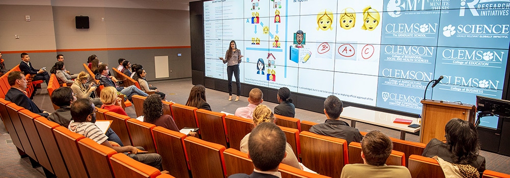 Woman on stage speaking to a crowd with monitors behind her showing emojis. 