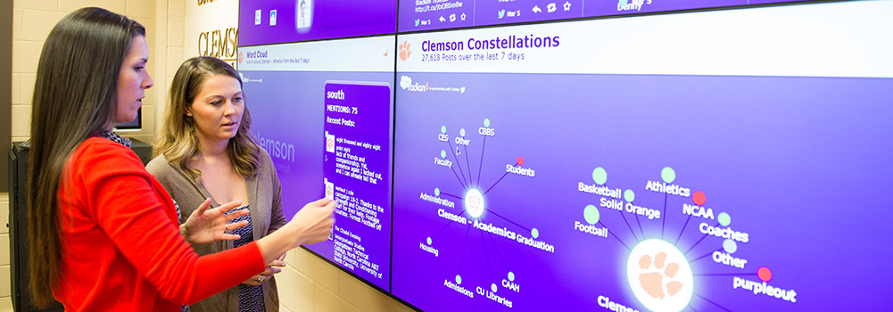 Two female students standing inside Clemson's Social Media Listening Center looking at monitors