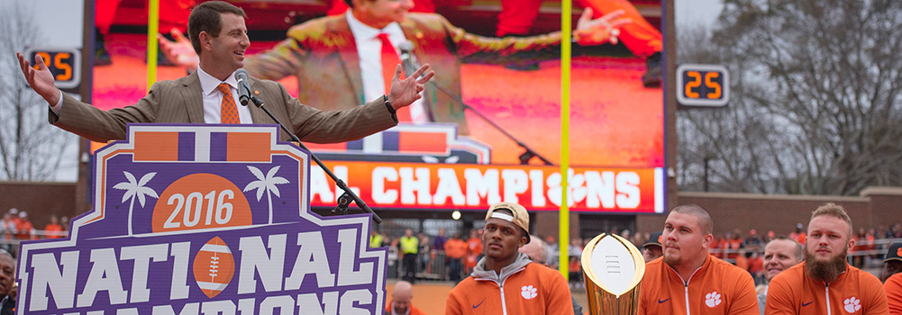 Football coach speaking at a podium to a crowd. Football players look on