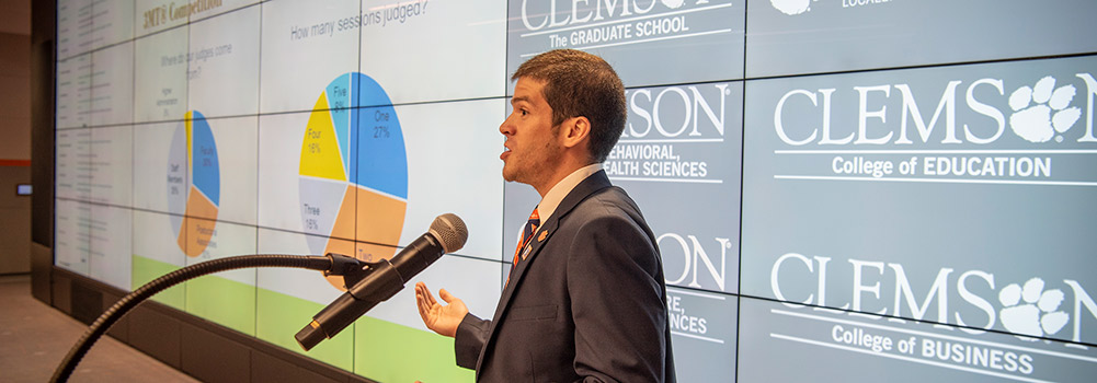 Man speaking to a crowd with indistinguishable pie charts on monitors in the background