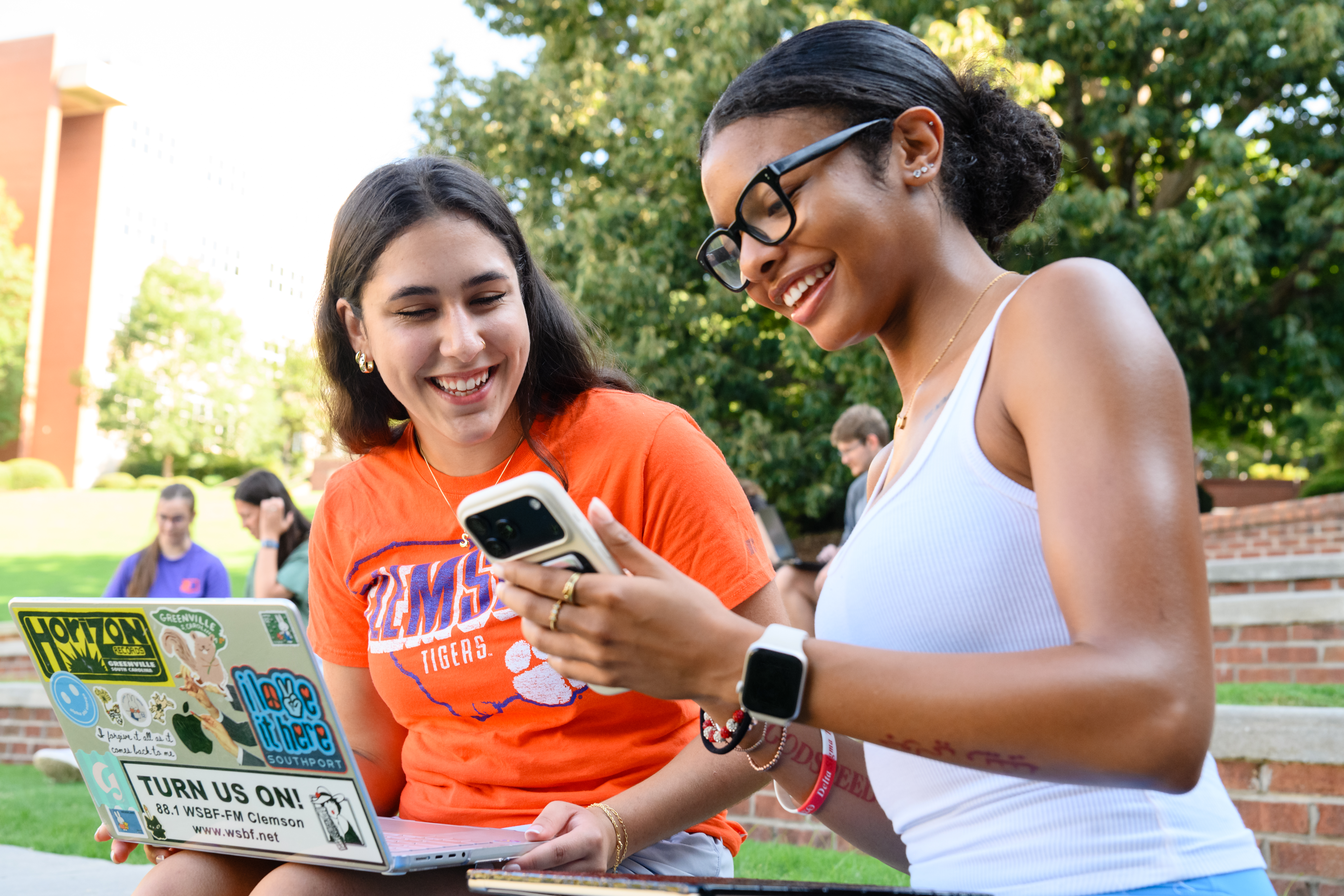 Two students outdoors at the amphitheater smile while looking at a smartphone, with a sticker-covered laptop open nearby.