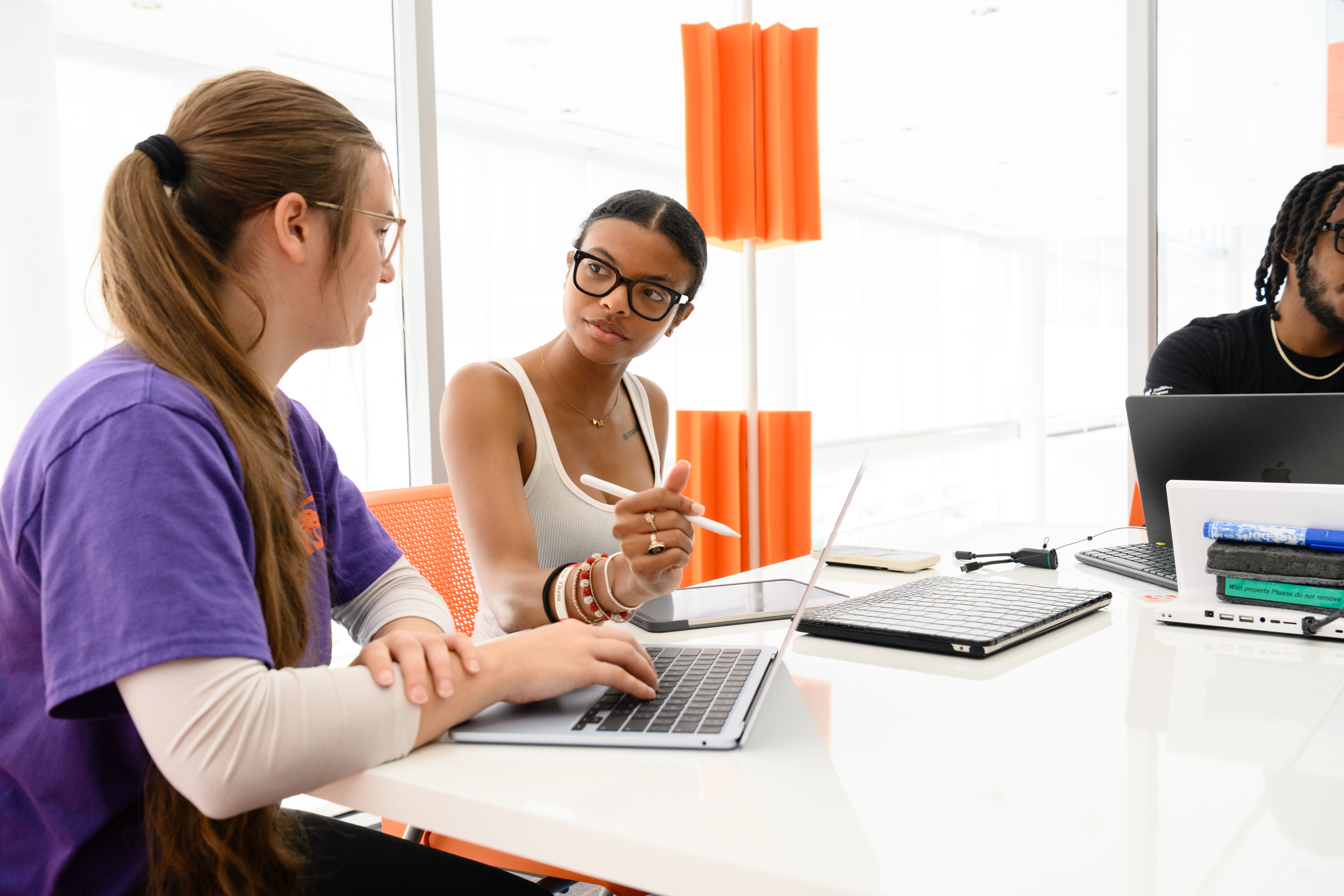 Three students collaborate at a table with laptops, with one person gesturing with a stylus while talking to another in a bright study space.