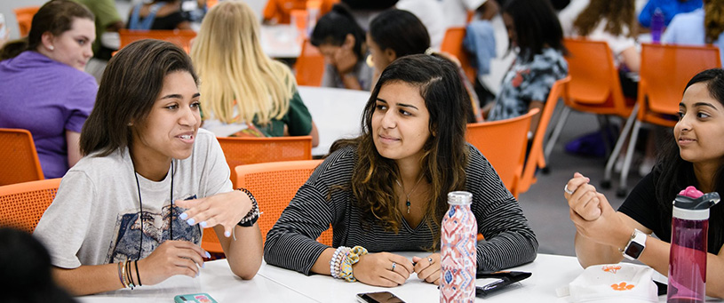 Three smiling young women sitting around a table listening to the person on the left while she talks. 