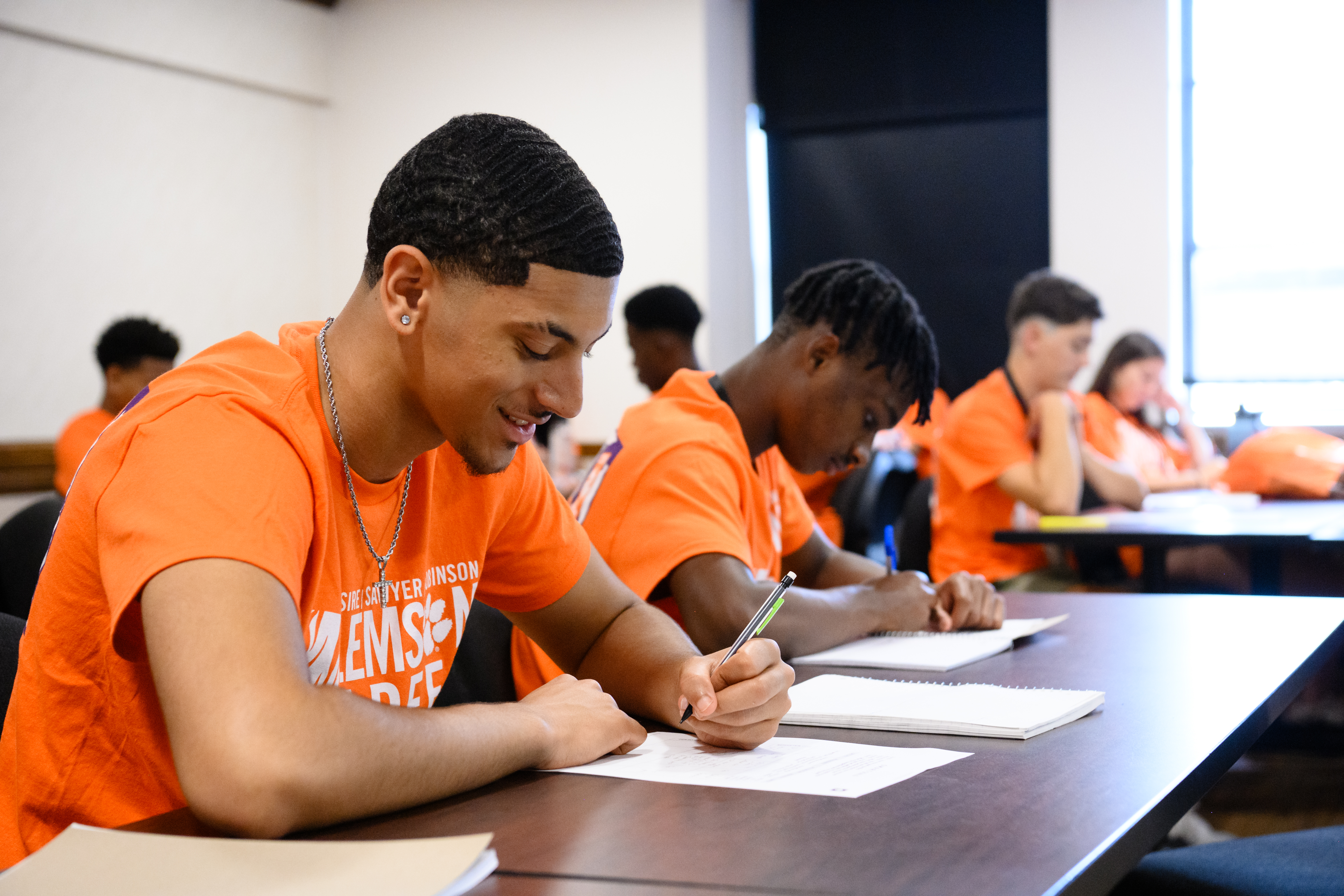 Students in matching orange T-shirts sit at classroom tables, taking notes and writing in notebooks.