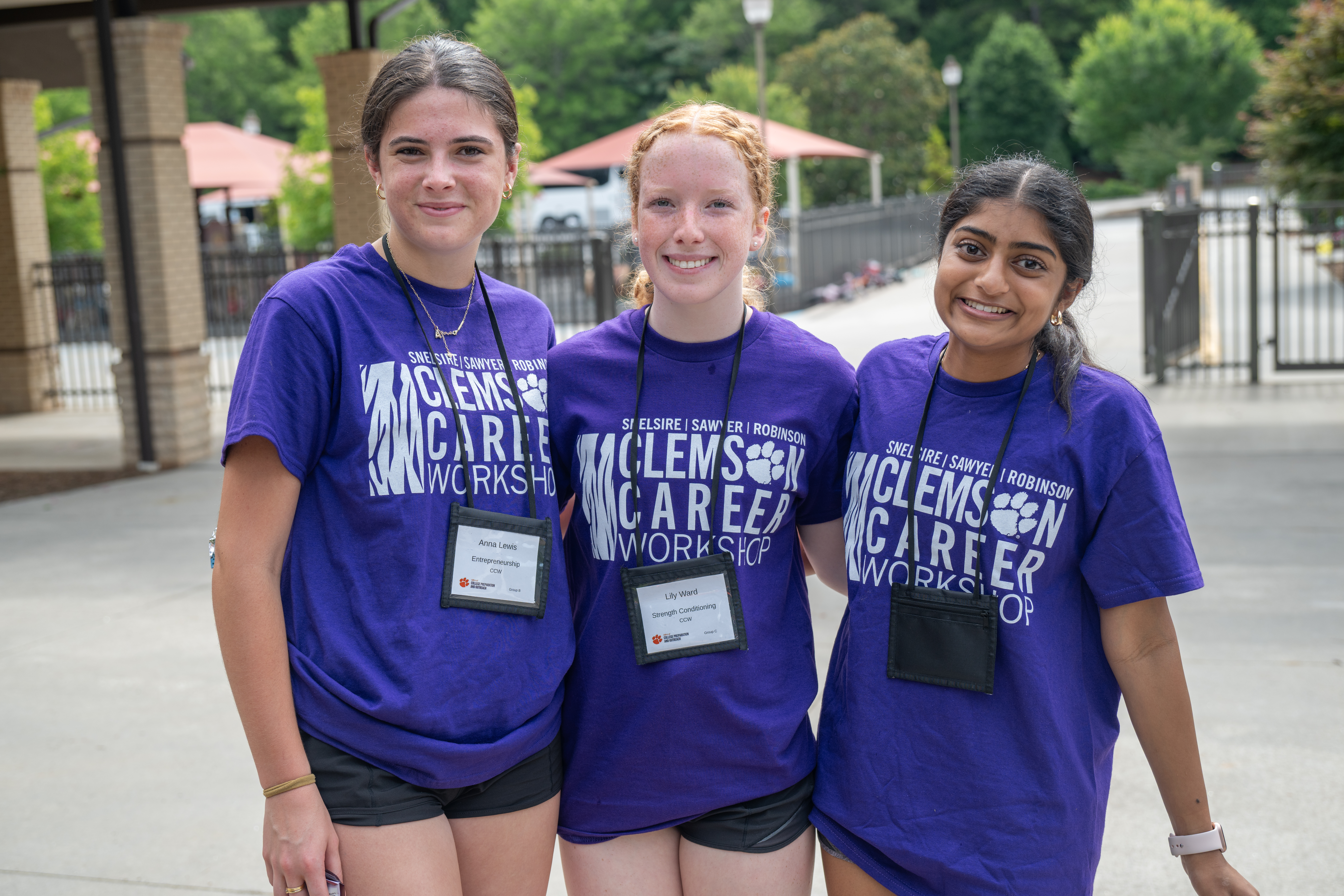 Three students in purple Clemson Career Workshop T-shirts pose together outdoors, wearing name badges on lanyards.