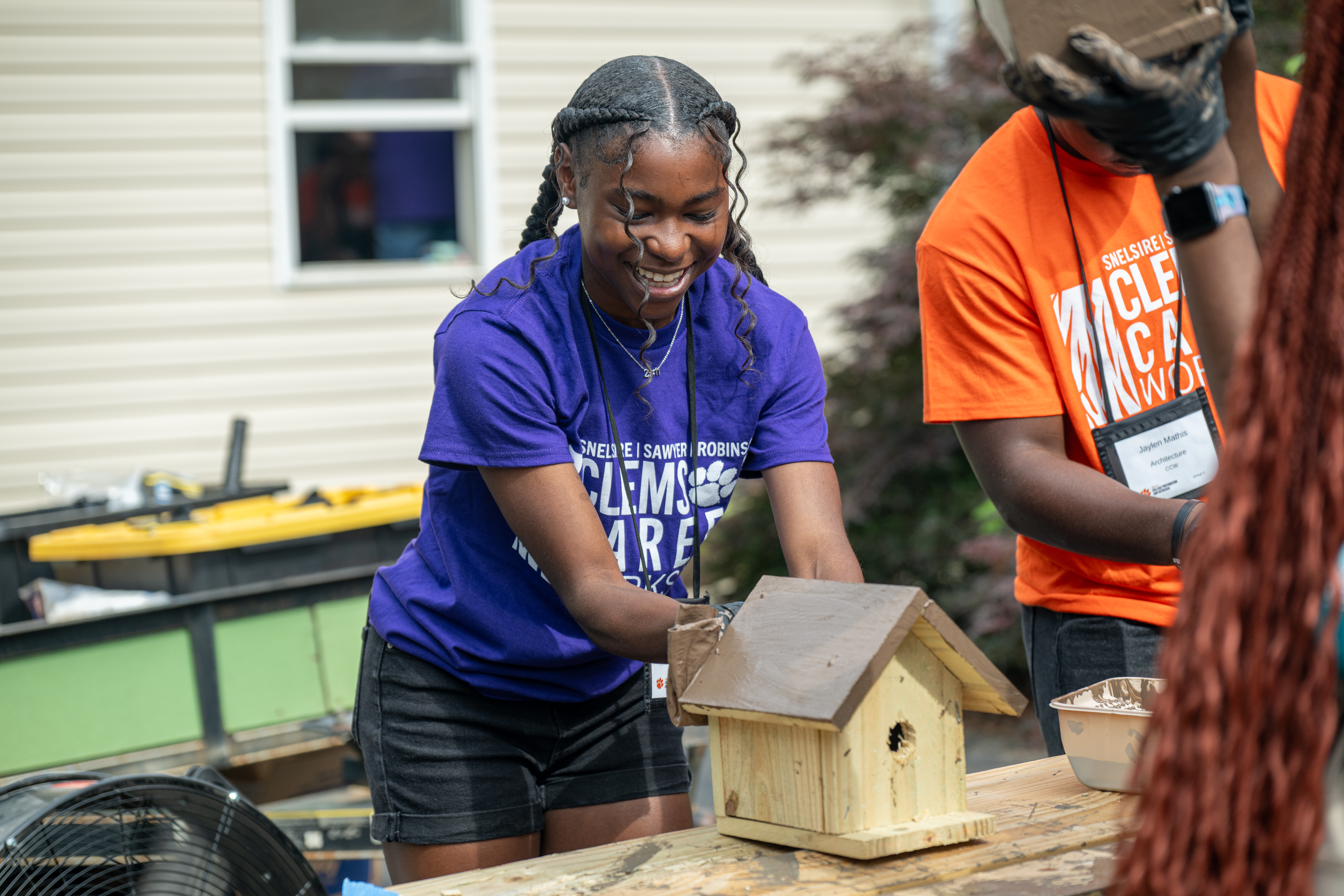 Smiling teenager paints a small wooden birdhouse at an outdoor worktable during a volunteer activity.