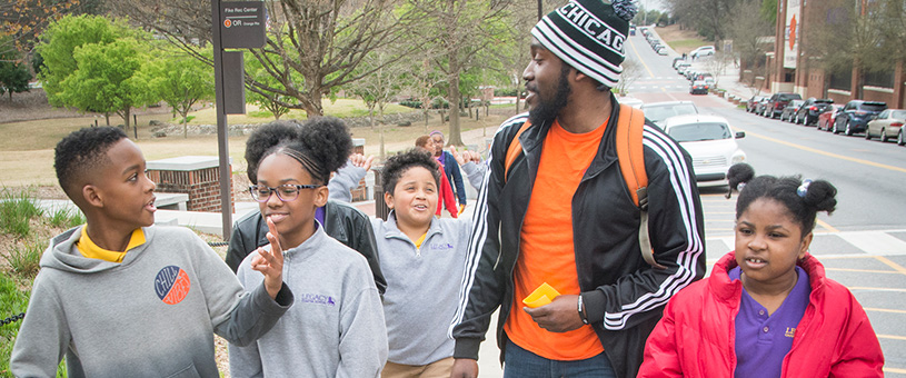 Group of middle school age kids walking on campus and talking excitedly. An adult leads the way as he looks back and smiles.