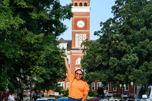 Student in orange, smiling as she raises her hand to showcase Clemson campus.