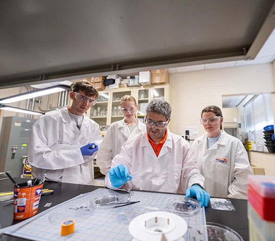 Several students gather around faculty member in the lab as he performs test.