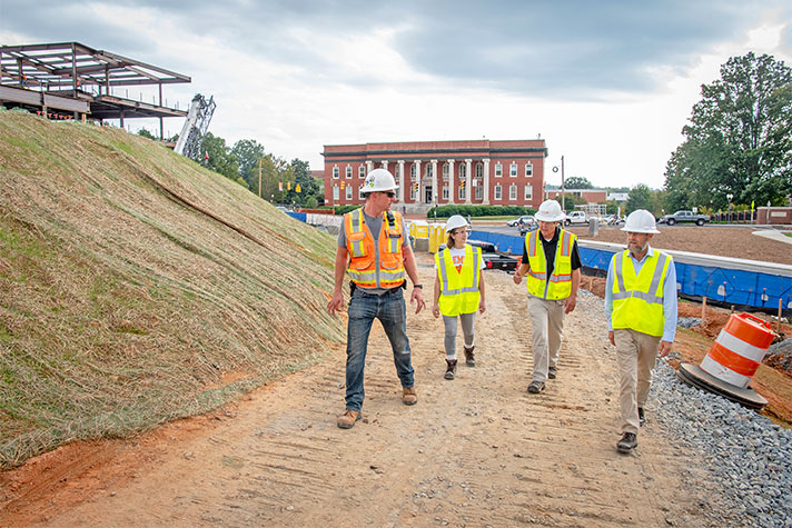Employees and student walk along construction site with Sikes Hall in the background.