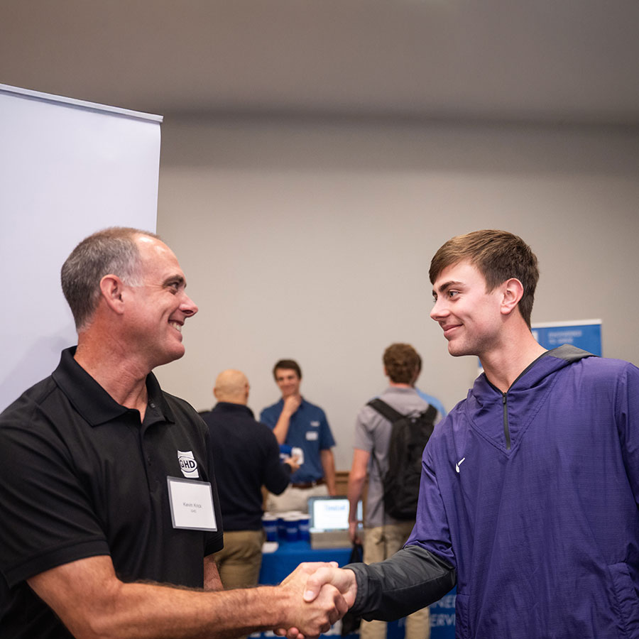 Industry professional and undergraduate student smiling at each other while they shake hands.