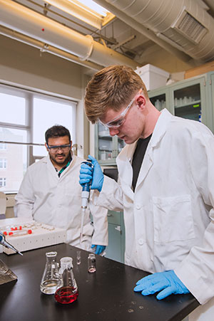 Male student using pipette with TA onlooking in lab