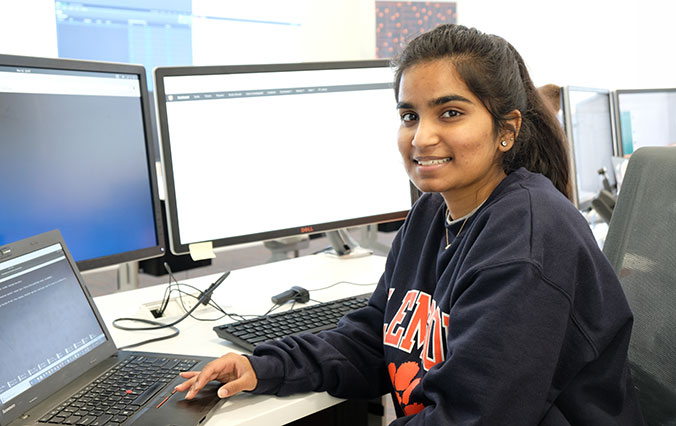 Female student wearing Clemson shirt while sitting in front of computer screens.