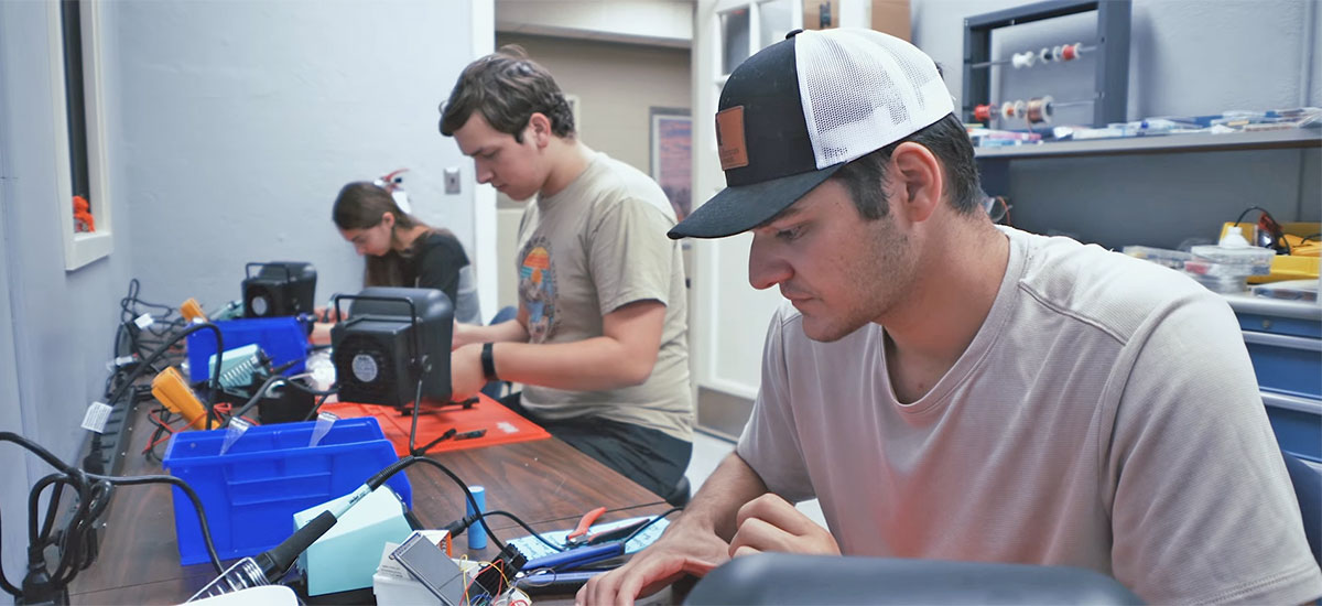 Three students are all focused, with their heads down, in the lab working on a project.