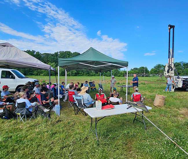 Students observing drilling demo in a field of grass.