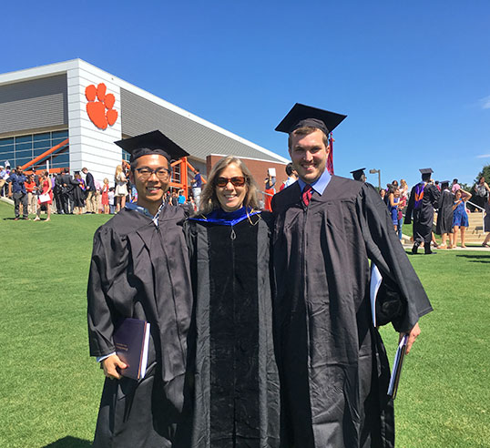 Graduates and faculty member stand together with Littlejohn in the background.