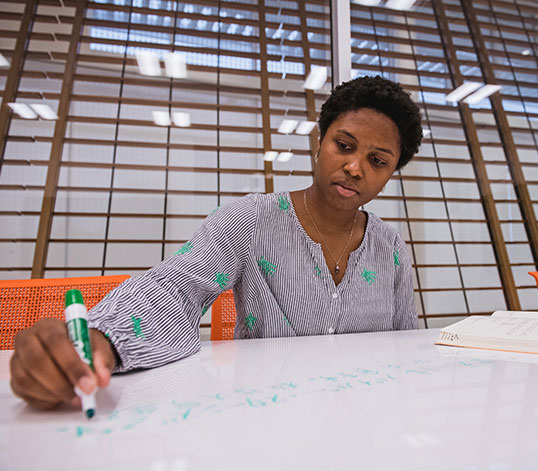 Student writing with book laying next to her on the table.
