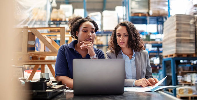 Two women on manufacturer floor viewing laptop.