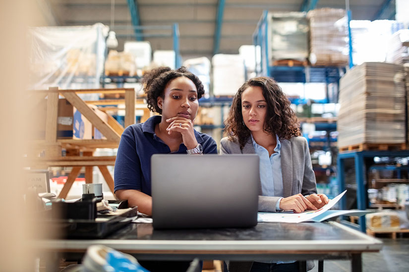 Two women looking at laptop on manufacturer floor.