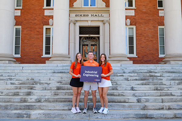 Three students in front of Sikes Hall holding up a sign that says Industrial Engineering.