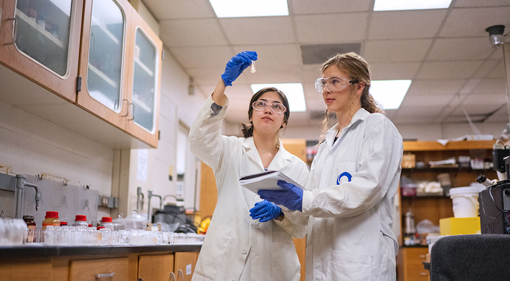 Two female students in lab looking up at tube.