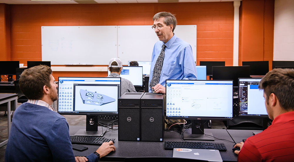 Students at computers in classroom with professor giving guidance.