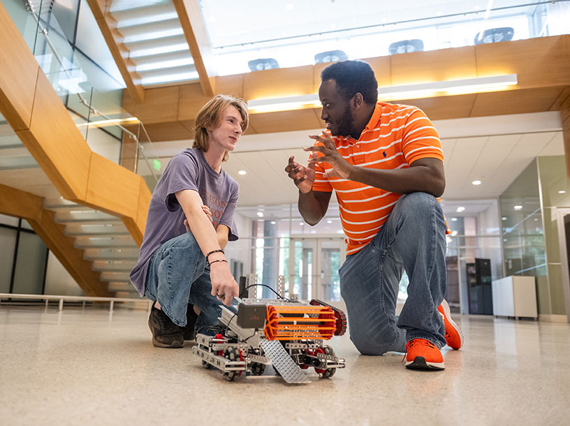 Two students talking, with small robotic vehicle situated in front of them.
