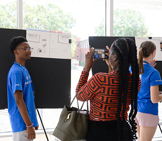 Participant posing in front of poster while his mother takes a photo.