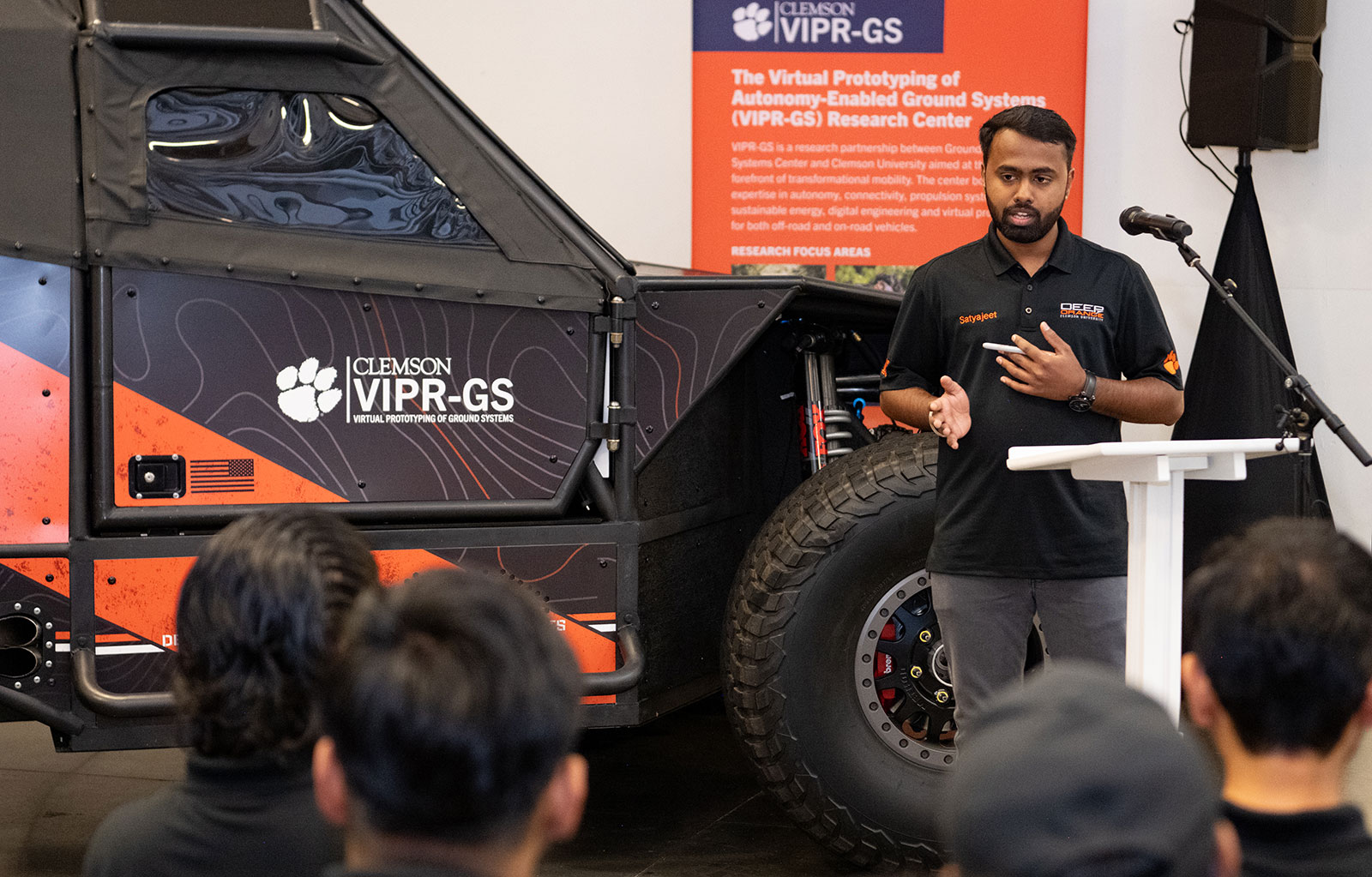 Students standing next to vehicle prototype he helped complete, while talking to a group about the work.