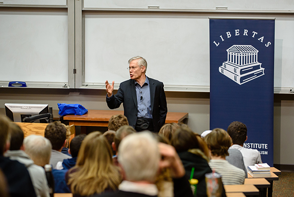 A speaker presenting to an audience at a lecture, with a banner displaying the "Libertas Institute" logo in the background.