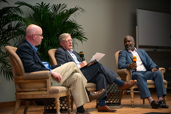 A panel discussion featuring three speakers in a comfortable setting, sitting in armchairs with microphones, engaging in conversation.